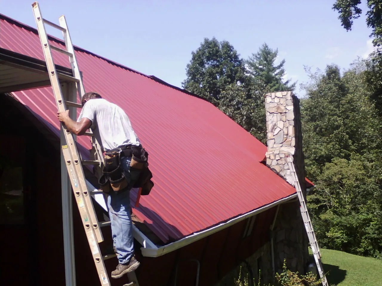 Roofer on a ladder beside a red metal roof and stone chimney — Soffit & Fascia Repair by Roofing Brothers in Clifton